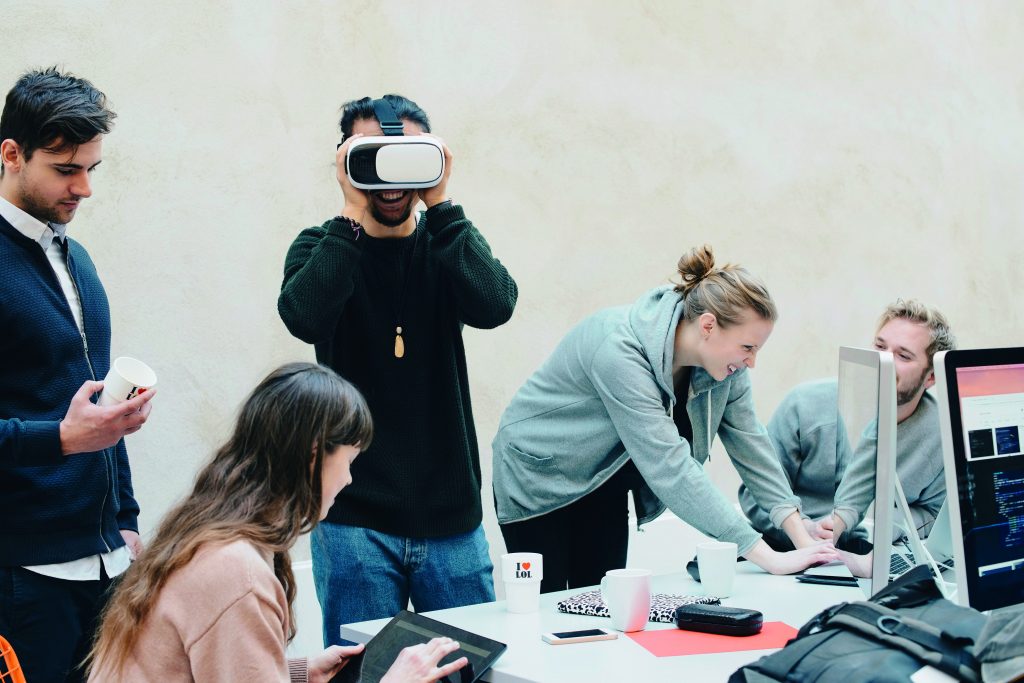 vijf jonge mensen rond een tafel met computers. Een ervan draagt een virtual-reality-bril; een ander werkt op een laptop; een meisje is bezig op een tablet