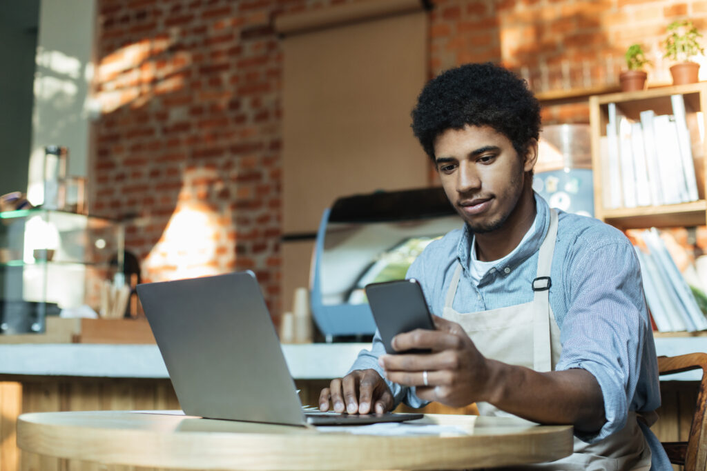 Een man zit achter een laptop en kijkt op zijn smartphone.