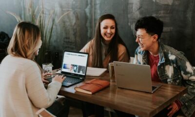 3 lachende mensen rondom een tafel. Er staan 2 laptops op en er liggen 2 notitieblokken.