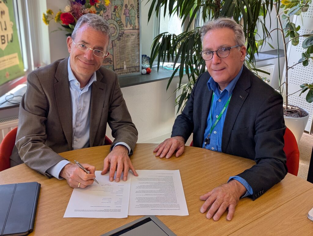 Twee mannen naast elkaar aan tafel, klaar om hun handtekening te zetten op de vellen papier voor ze.