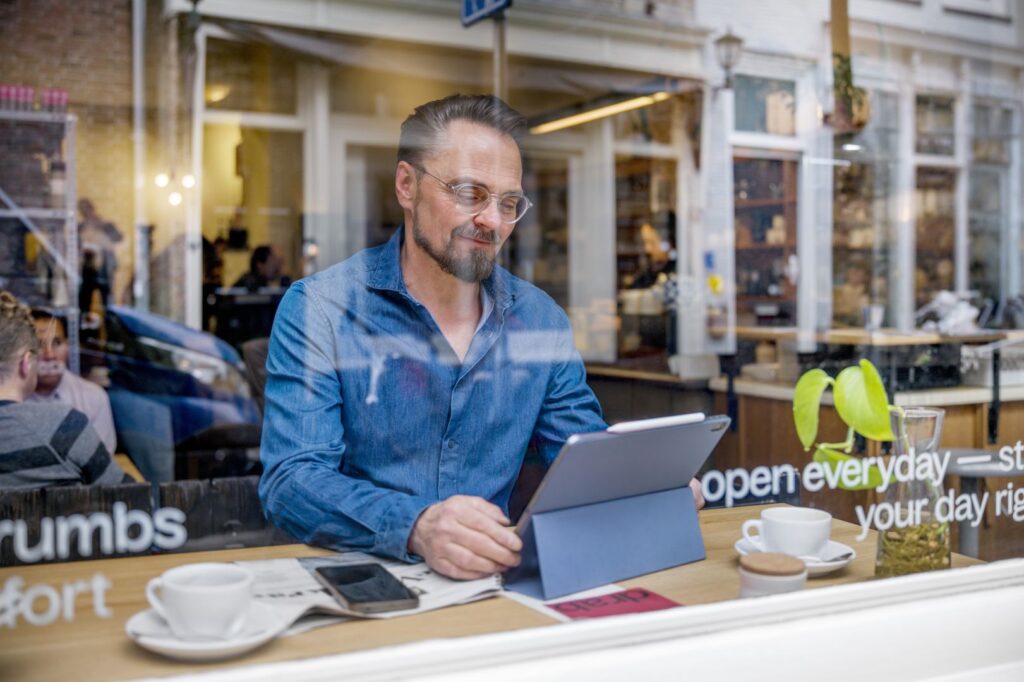 Man in café op laptop aan tafel bij het raam; foto van buiten genomen. 