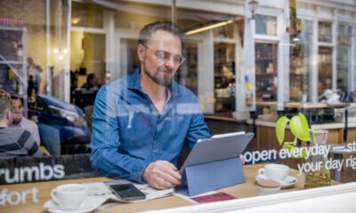 Man in café op laptop aan tafel bij het raam; foto van buiten genomen.